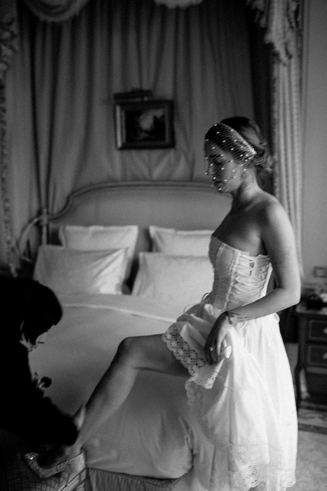 Bride in a pearl birdcage veil fitted for her shoe beside a canopied bed at the Ritz, photographed by Thierry Joubert