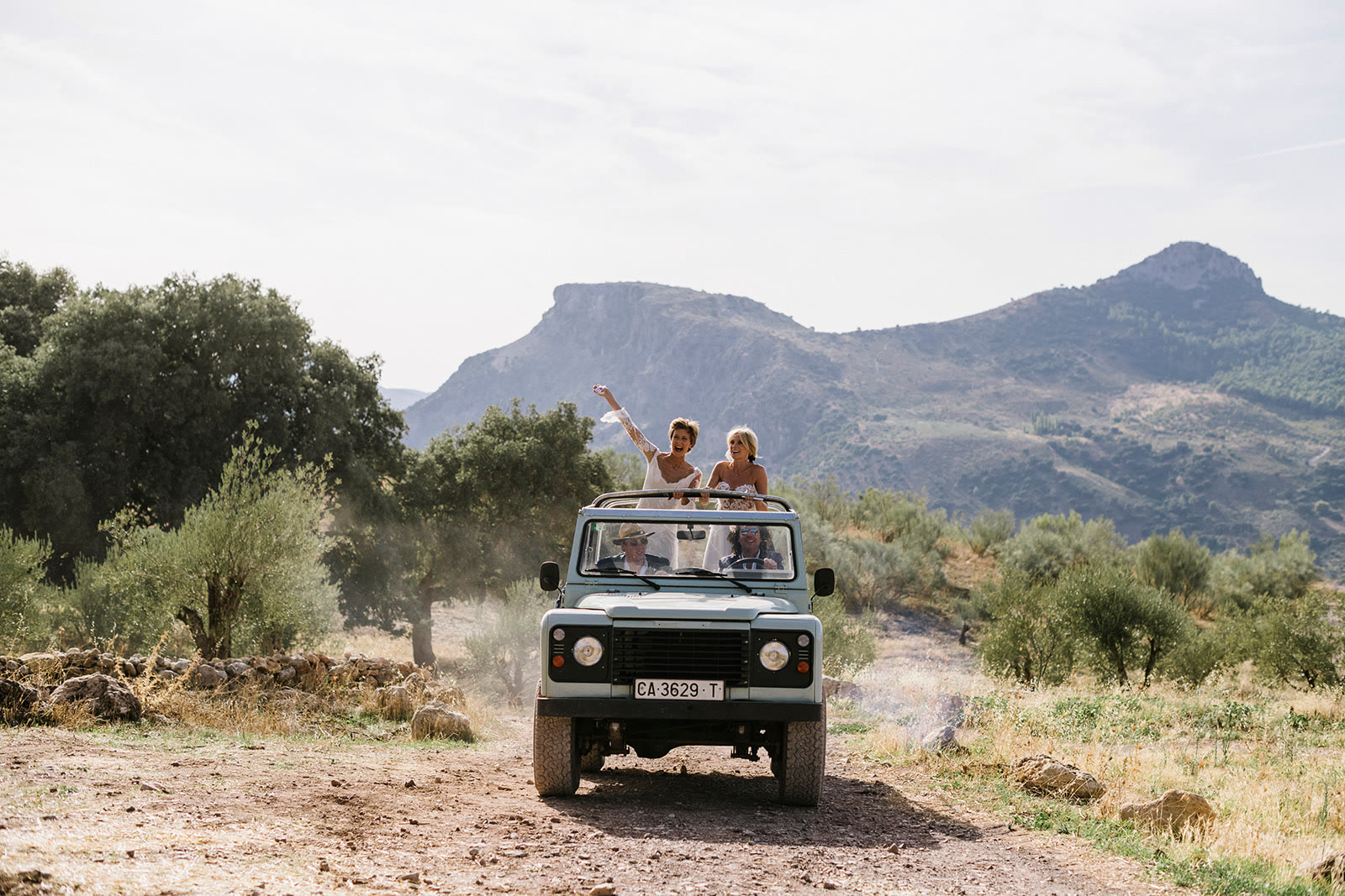 Two brides cheering from an open-top vintage vehicle, photographed by Thierry Joubert